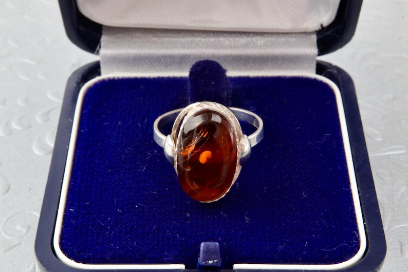 Puede incluir: Un anillo de plata con una piedra de &aacute;mbar de forma ovalada engastada en un engaste de plata. El anillo est&aacute; en una caja de anillo de terciopelo azul.