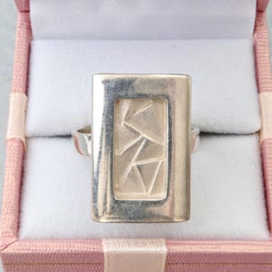 Op de afbeelding: Een zilveren ring met een groot, rechthoekig oppervlak met een gladde, gepolijste rand rondom een verdiept middenpaneel. Het middenpaneel heeft een getextureerde, matte afwerking met een reeks ingesneden, hoekige geometrische lijnen.