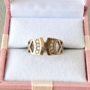 Puede incluir: Un anillo dorado con un diseño geométrico, con una sección central con seis pequeñas piedras transparentes. El anillo tiene calados en forma de diamante en ambos lados. Se presenta dentro de un joyero rosa con interior blanco.