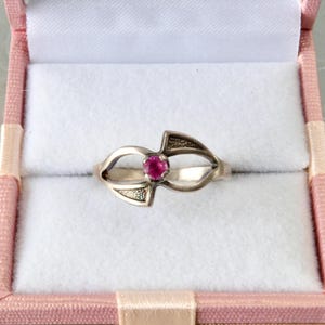 May include: A silver ring with a central, round, red gemstone. The ring has an open design with decorative elements on either side of the gemstone. The ring is displayed inside a pink jewelry box with a white interior.