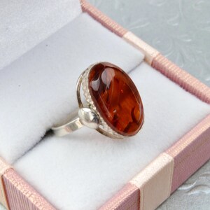 Puede incluir: Un anillo de plata con una piedra de &aacute;mbar de forma ovalada engastada en un engaste de plata. El anillo est&aacute; en una caja de regalo rosa y blanca.