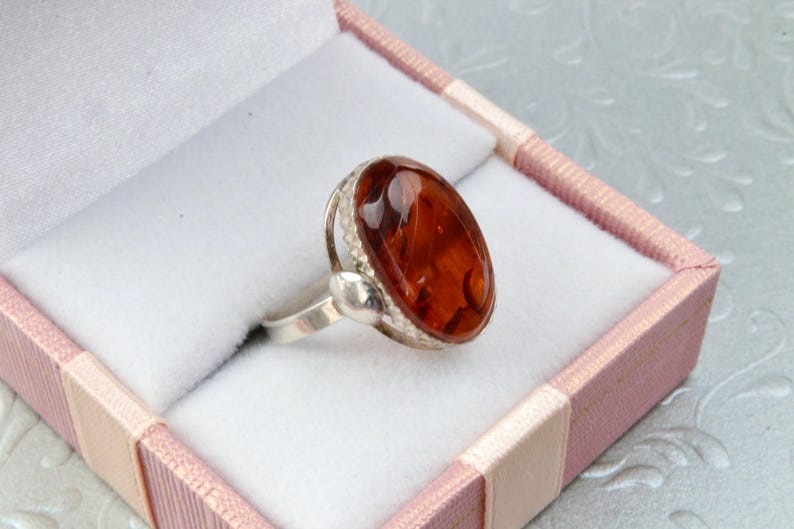 Puede incluir: Un anillo de plata con una piedra de &aacute;mbar de forma ovalada engastada en un engaste de plata. El anillo est&aacute; en una caja de regalo rosa y blanca.