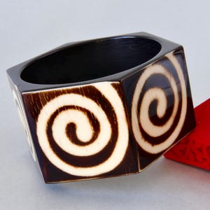 Könnte beinhalten: Ein sechseckiges Armreif-Armband mit dunkelbrauner Außenseite und cremefarbenen Spiralmustern. Die Innenseite ist schwarz. Eine rote Schachtel ist im Hintergrund zu sehen. Dieses einzigartige Armband ist ein Modeaccessoire.