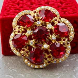 Può includere: Spilla dorata a forma di fiore con grandi gemme rosse sfaccettate e cristalli trasparenti più piccoli. La spilla è appoggiata su un cofanetto di velluto rosso. Gioiello vintage.