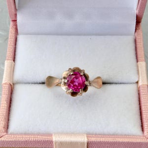 Op de afbeelding: Een goudkleurige ring met een helder roze edelsteen, gezet in een bloemmotief. De ring wordt gepresenteerd in een roze sieradendoosje met een witte binnenkant. Het ontwerp van de ring is vintage.