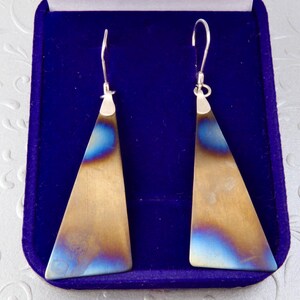 Peut inclure: Une paire de boucles d'oreilles pendantes composées de plaques métalliques triangulaires allongées à la finition brossée. Le métal présente un dégradé de tons or métallique et bleu irisé. Chaque boucle est fixée à un crochet argenté.