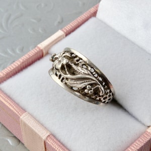 Op de afbeelding: Een zilveren ring met een sierlijk bloemmotief, gepresenteerd in een roze en witte sieradendoos. De ring heeft ingewikkeld filigraanwerk en kleine, ronde versieringen. De ring is een sieraad.