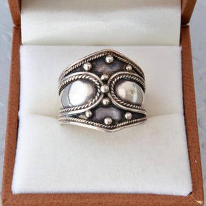 Può includere: Un anello color argento con un design ornato, caratterizzato da due forme ovali e piccoli accenti rotondi. L'anello è presentato in un cofanetto marrone con interno bianco, che mette in risalto i dettagli intricati dell'anello.
