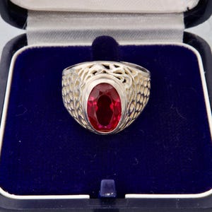 Op de afbeelding: Een zilveren ring met een ovale, dieprode edelsteen, gezet in een openwerk ontwerp. De ring wordt gepresenteerd in een blauw fluwelen sieradendoosje met een donkerblauwe buitenkant.