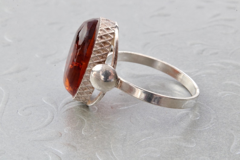 Puede incluir: Un anillo de plata con una gran piedra preciosa ovalada de &aacute;mbar. El anillo tiene una banda texturizada y un engaste redondo y liso para la piedra.