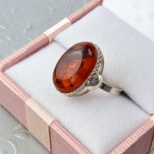 Puede incluir: Un anillo de plata con una gran piedra ovalada de &aacute;mbar engastada en un bisel decorativo. El anillo se muestra en una caja de anillo de terciopelo rosa.