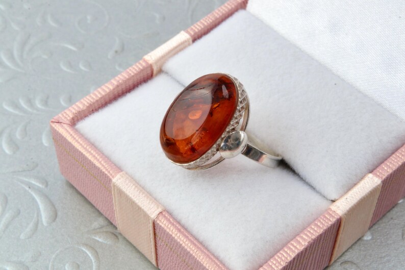 Puede incluir: Un anillo de plata con una gran piedra ovalada de &aacute;mbar engastada en un bisel decorativo. El anillo se muestra en una caja de anillo de terciopelo rosa.