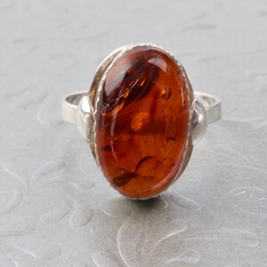 Puede incluir: Un anillo de plata con una piedra preciosa de &aacute;mbar de forma ovalada. El &aacute;mbar es de color naranja oscuro con inclusiones visibles.