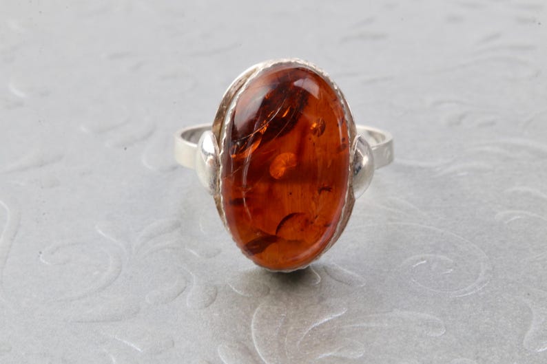 Puede incluir: Un anillo de plata con una piedra preciosa de &aacute;mbar de forma ovalada. El &aacute;mbar es de color naranja oscuro con inclusiones visibles.