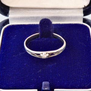 May include: A silver-toned ring featuring a small, central flower-shaped setting with a single clear stone. The band has an open, filigree-style design on both sides of the centre. The ring is displayed inside a dark blue velvet-lined jewellery box.