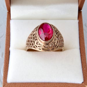 Può includere: Un anello in oro con una grande pietra preziosa di colore rosso vibrante dal taglio ovale, incastonata in una lunetta centrale. La fascia presenta un intricato design in filigrana traforata che avvolge il dito con un motivo metallico.