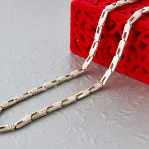 Puede incluir: Un collar plateado con un diseño de eslabones rectangulares. El collar se exhibe sobre una superficie gris claro junto a una caja roja cubierta de terciopelo con un patrón decorativo.