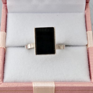 Può includere: Un anello d'argento con una pietra nera rettangolare, presentato in un cofanetto rosa con interno bianco. La fascia dell'anello è sottile e semplice, che completa la pietra scura e audace. La scatola è chiusa, suggerendo un regalo o un deposito.