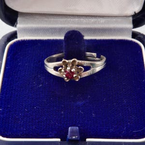 Puede incluir: Un anillo de plata con un diseño en forma de flor y una gema roja en el centro, exhibido en una caja de joyería de terciopelo azul. El anillo tiene un diseño de banda dividida. La caja está abierta.