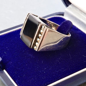 Puede incluir: Un anillo plateado con una gran piedra negra rectangular. El anillo está engastado en una banda plateada con pequeños detalles decorativos. Presentado en un joyero de terciopelo azul, de estilo vintage.