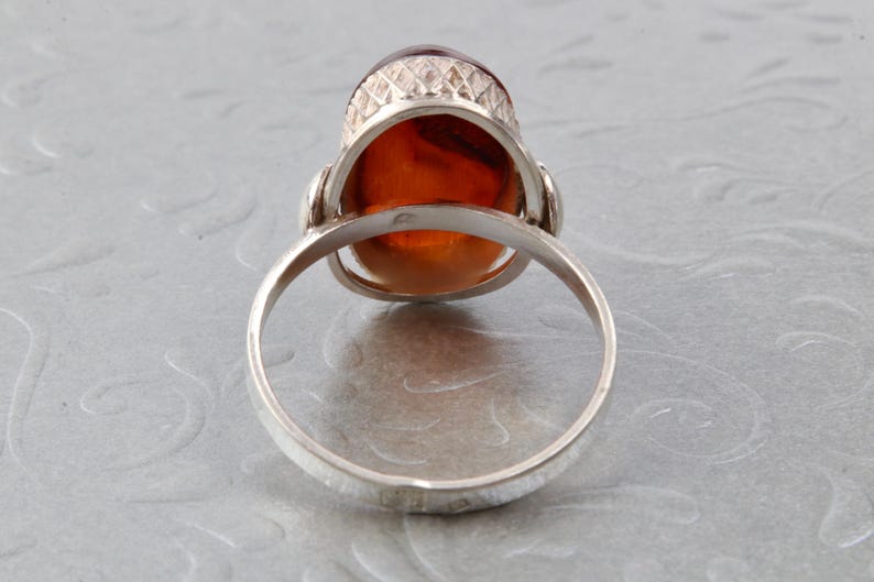 Puede incluir: Un anillo de plata con una piedra de &aacute;mbar grande de forma ovalada engastada en un engaste decorativo. El anillo se muestra desde atr&aacute;s, revelando la banda lisa.