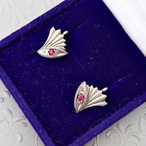 Può includere: Un paio di orecchini color argento con un design a ventaglio, ciascuno con una piccola gemma rossa rotonda. Gli orecchini sono esposti in un portagioie di velluto blu con fodera di raso bianco.