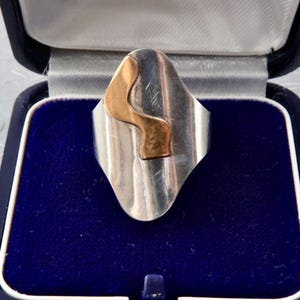 Könnte beinhalten: Silberfarbener Ring mit goldfarbenem, abstraktem Design. Der Ring befindet sich in einer blauen Samt-Schmuckschatulle mit dunkelblauem Äußeren. Das Design des Rings ist eine geschwungene, längliche Form.