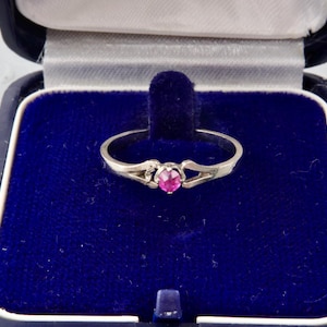 Può includere: Un anello d'argento con una piccola gemma rosa rotonda al centro. L'anello è presentato in un portagioie di velluto blu con fodera in raso bianco. Il design dell'anello è delicato e aperto.