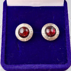 Può includere: Un paio di gemelli rotondi con un bordo color argento e un motivo a chiave greca. Ogni gemello ha una pietra centrale circolare rosso intenso. I gemelli sono esposti in un cofanetto di velluto blu.