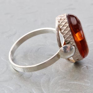 Puede incluir: Un anillo de plata con una piedra de &aacute;mbar grande de forma ovalada. La piedra est&aacute; engastada en un engaste de plata con un dise&ntilde;o texturizado. La banda del anillo es lisa y sencilla.