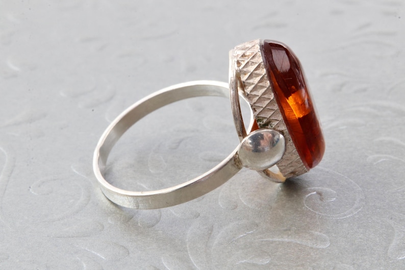 Puede incluir: Un anillo de plata con una piedra de &aacute;mbar grande de forma ovalada. La piedra est&aacute; engastada en un engaste de plata con un dise&ntilde;o texturizado. La banda del anillo es lisa y sencilla.