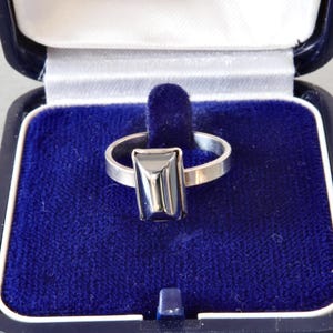 Puede incluir: Un anillo de metal plateado con una sola piedra rectangular facetada de color gris oscuro montada verticalmente. El anillo se muestra dentro de un joyero forrado de terciopelo azul oscuro con un forro de satén blanco.