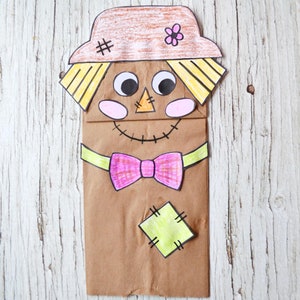 May include: A handmade scarecrow puppet made from a brown paper bag. The scarecrow has a tan hat with a flower and stitches, large googly eyes, a triangle nose, and a stitched smile. A pink bow tie and a green patch complete the design.