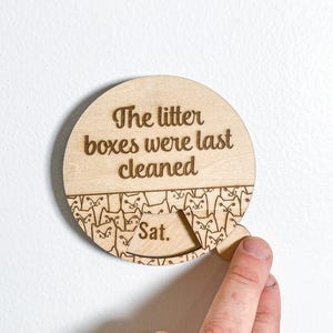 May include: A circular light-colored wooden tracker with the engraved text "The litter boxes were last cleaned" above a pattern of cat faces. A small sliding wooden tab reveals the day of the week, currently displaying the abbreviation "Sat."
