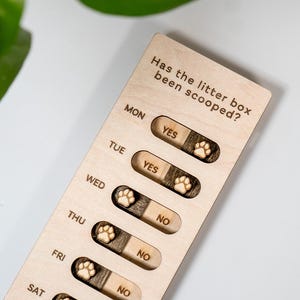 May include: A wooden cat litter box tracker with the text "Has the litter box been scooped?" and the days of the week. Each day has a sliding indicator with "Yes" or "No" options and a paw print design.