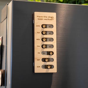 May include: A wooden dog walking tracker with the text "Have the dogs been walked?" and days of the week. Each day has a paw print icon and a "Yes" or "No" slider. The tracker is attached to a dark gray refrigerator.
