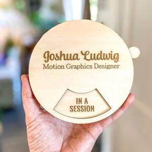 May include: A round wooden sign with the name "Joshua Ludwig" and the title "Motion Graphics Designer" engraved on it. The sign also features the text "IN A SESSION" in a cut-out section.