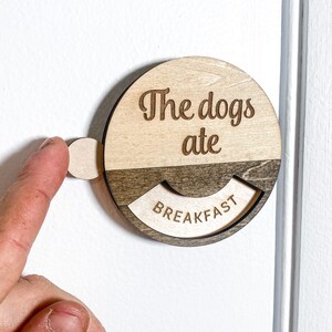 May include: Wooden sign with the text "The dogs ate" above a rotating section that reads "BREAKFAST". The sign is round with a light wood finish and a darker wood accent. A small, round tab extends from the side for turning.