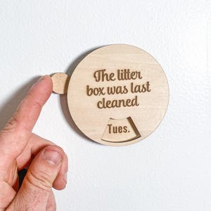 Peut inclure: Un traqueur circulaire en bois clair avec le texte gravé "The litter box was last cleaned" et une petite fenêtre découpée affichant le jour "Tues." Le dispositif comporte un petit bouton circulaire sur le côté gauche pour régler le jour.