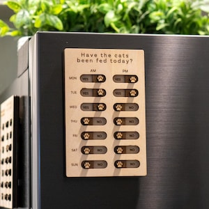Könnte beinhalten: Ein hölzerner Katzenfutter-Tracker mit dem Text "Have the cats been fed today?" und einem Raster mit den Wochentagen sowie den Spalten "AM" und "PM". Jeder Tag hat eine Option "YES" und "NO" mit einem Pfotenabdruck-Symbol.