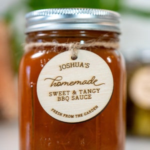 Puede incluir: Un tarro de vidrio lleno de salsa marrón, etiquetado con una etiqueta redonda de madera que dice "Joshua's Homemade Sweet & Tangy BBQ Sauce Fresh From The Garden".