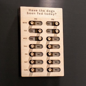 Op de afbeelding: Houten hondenvoederschema met de tekst "Have the dogs been fed today?" en pootafdrukindicatoren. De kaart heeft tuimelschakelaars voor AM en PM, en dagen van de week. De kaart is bevestigd aan een zwarte koelkast.
