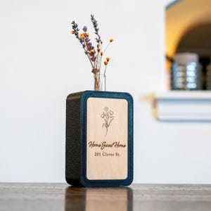 Pode incluir: Um vaso retangular com uma moldura azul escuro e um painel de madeira clara. O painel apresenta um design floral e o texto "Home Sweet Home, 201 Clover St." Um pequeno frasco de vidro contém flores secas.