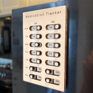 Pode incluir: Um rastreador de medicamentos retangular de madeira com o texto Medication Tracker no topo. Possui colunas para AM e PM e linhas para cada dia da semana. Cada linha tem um interruptor deslizante com etiquetas YES e NO para doses.
