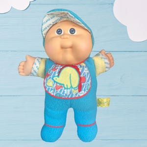 May include: A Cabbage Patch Kid doll wearing a blue and yellow outfit with a dinosaur print. The doll is wearing a blue hat with the words "Babyland Kid" printed on it.