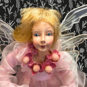 May include: A detailed close-up of a fairy doll with blonde hair and blue eyes. The doll wears a pink dress with a matching floral collar. The fairy has translucent wings and is set against a black background with a white floral pattern.