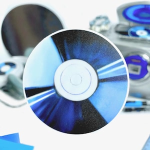 Puede incluir: Un disco compacto azul y negro con un centro plateado. El disco está sobre una superficie blanca.