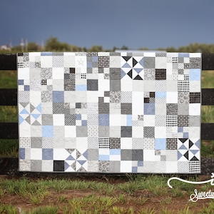 Puede incluir: Una colcha de patchwork con una cuadrícula de bloques de tela cuadrados en tonos negro, blanco, gris y azul claro. Los patrones incluyen rayas, puntos, estrellas y formas geométricas. El texto "ink Sweetwater" está impreso en blanco.