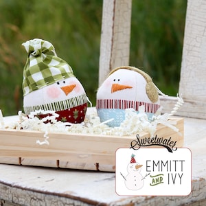 May include: Two snowman-shaped fabric ornaments. One wears a green and white checkered hat and a red and green patterned bottom. The other has earmuffs and a blue and white patterned bottom. Both have orange carrot noses and black dot eyes.
