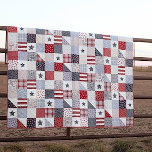 May include: A red, white, and blue patchwork quilt with stars and stripes. The quilt is hanging on a wooden fence.